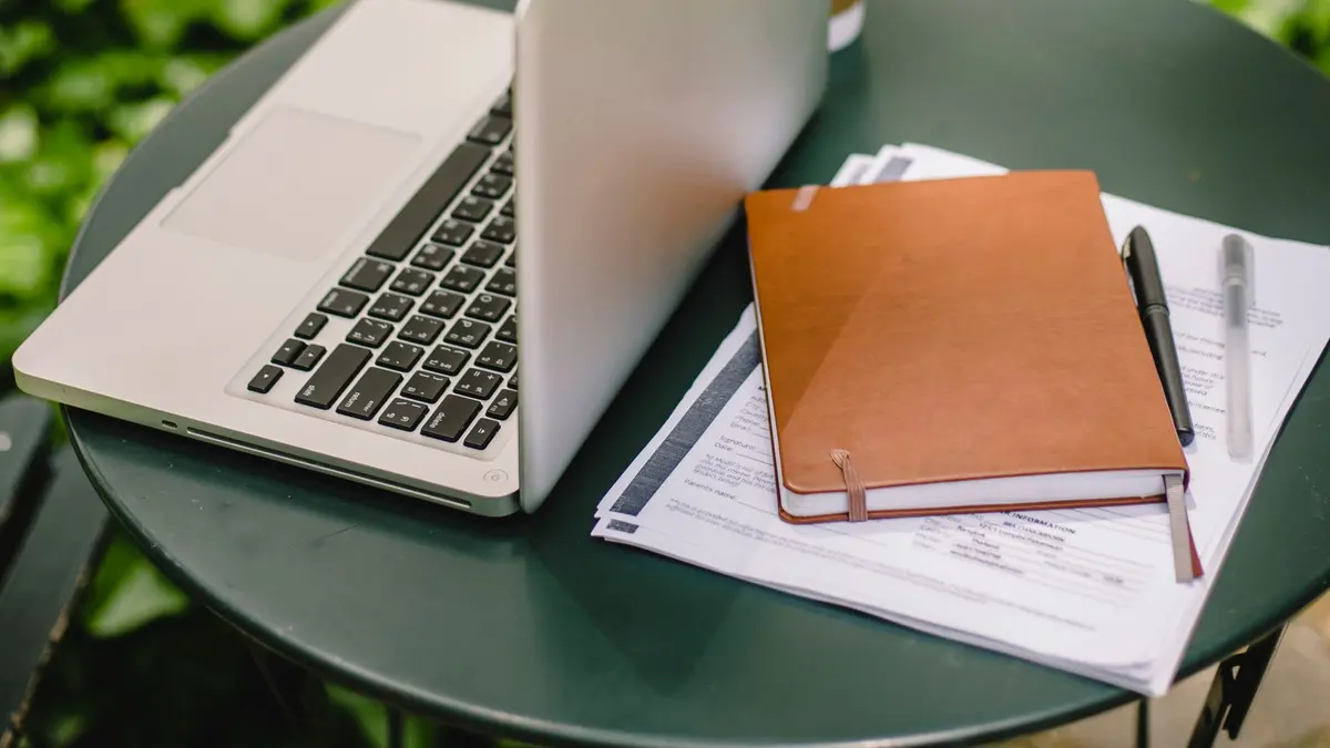 A laptop beside a stack of student visa application forms, a notebook and a pen on a table